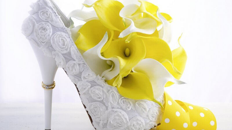 Trauring hängt am Absatz eines weißen hohen Brautschuhs mit gelben Blüten und Schleife
