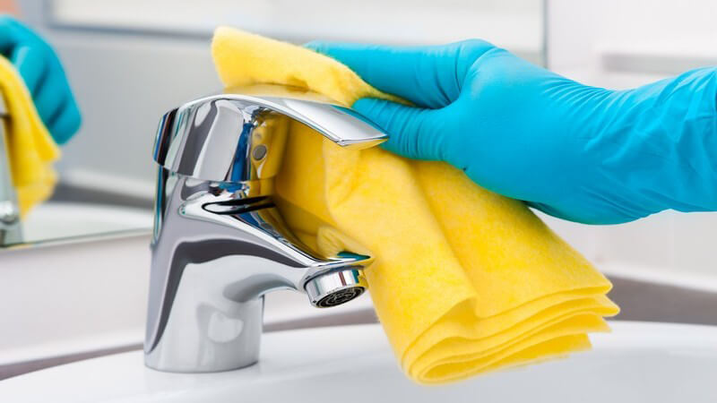Putzen einer Badarmatur mit gelbem Lappen und blauen Handschuhen