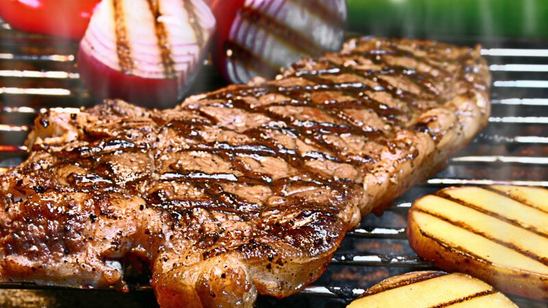 Fertiges Steak auf dem Grill, daneben gegrillte Kartoffelscheiben und Zwiebeln