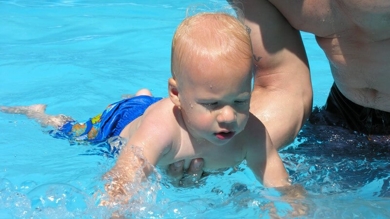 Blondes Baby in blauer Schwimmhose in Schwimmbecken, wird von Vater mit rechtem Arm gehalten, Freibad