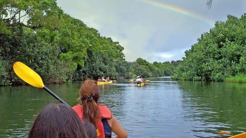 Kanu fährt durchs Wasser, davor fahren zwei Kanus, links und rechts Bäume, am Himmel Regenbogen