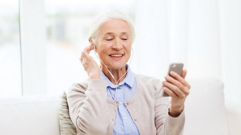 Seniorin in Strickjacke sitzt mit Smartphone und Kopfhörern auf der Couch
