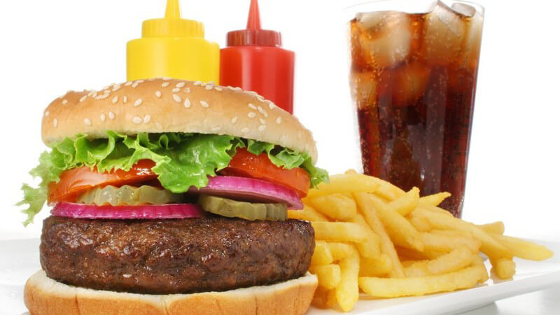 Hamburger mit Pommes, dahinter Ketchup, Mayonnaise und ein Glas mit Cola