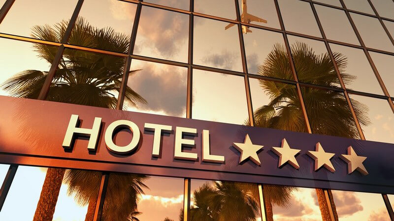 Blick auf die Front eines Hotels mit vier Sternen, im Gebäude spiegeln sich Palmen und ein Flugzeug