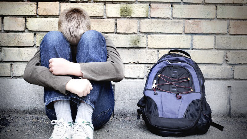 Trauriger Junge sitzt zusammengekauert an Wand, einsam, mit Schulrucksack neben sich