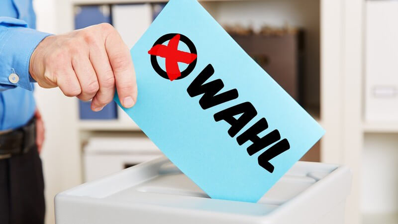 Mann steckt einen Stimmzettel mit der Aufschrift "WAHL" und einem roten Kreuz in die Wahlurne