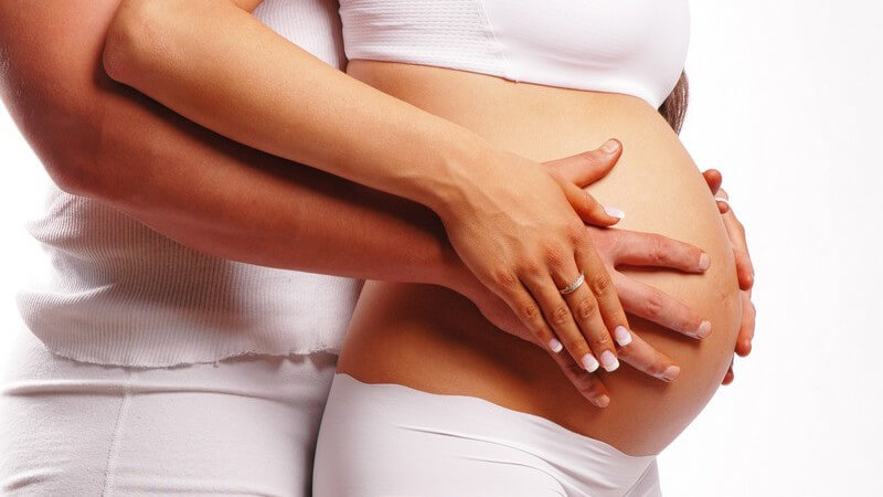 Schwangerer Bauch im Profil, die Hände der Eltern streichen zum Bauchnabel hin, beide Personen in weiß