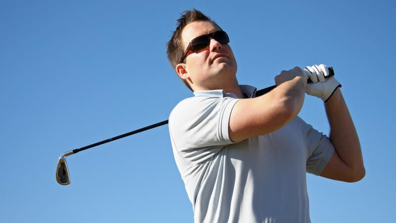 Oberkörper von Mann mit Golfhandschuhen und Sonnenbrille nach Abschlag mit Golfschläger über der Schulter, Himmel
