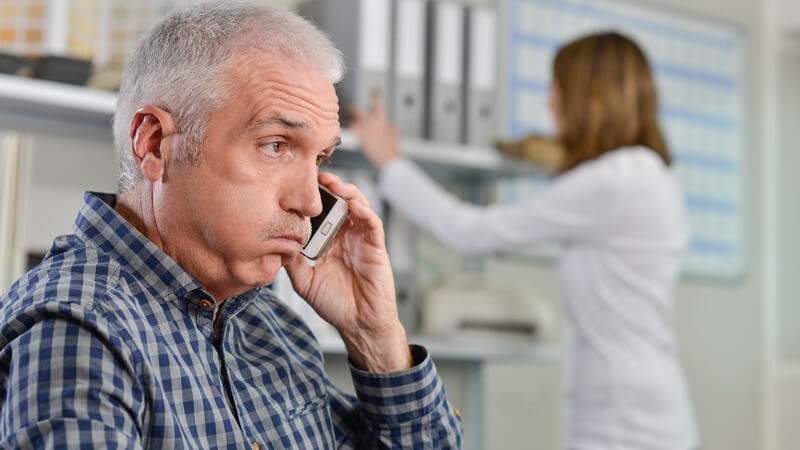 Mann mit grauen Haaren telefoniert im Büro mit dem Smartphone und guckt gestresst, im Hintergrund steht eine Kollegin