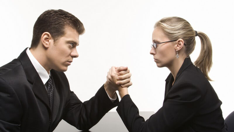 Mann und Frau im Businessoutfit schauen sich kalt an und machen Armdrücken