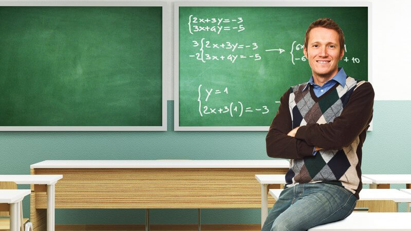 Junger Lehrer sitzt auf Tisch in Klassenzimmer, im Hintergrund Tafel mit Matheformeln