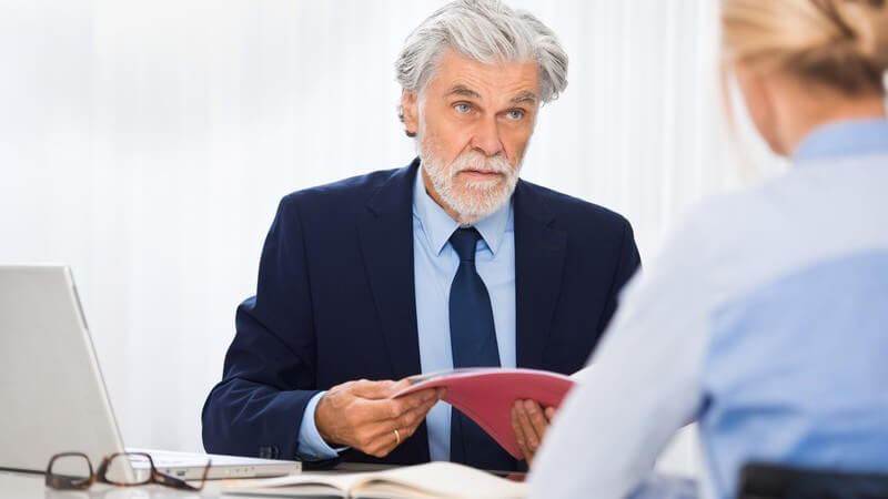 Älterer Chef mit grauen Haaren und Vollbart sitzt einer blonden Bewerberin gegenüber und betrachtet die Bewerbungsmappe