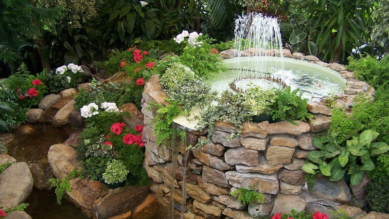 Springbrunnen aus Stein mit Wasserspiel im Garten, umgeben von Pflanzen