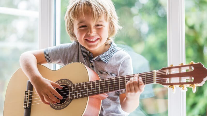 Blonder Junge sitzt an einem Fenster und spielt Akustikgitarre