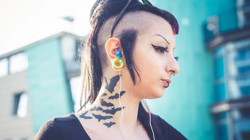 Junge Frau im Gothic-Stil, abrasierte Kopfhaare an der Seite, Fledermaus-Tattoo am Hals und diverse Piercings