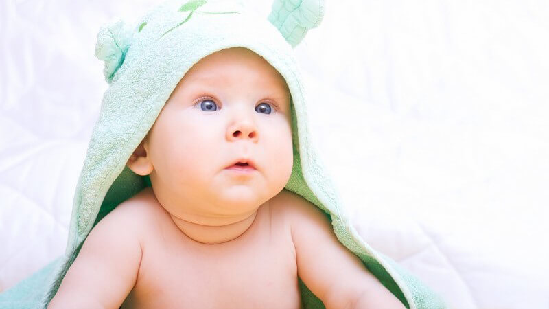 Junges Baby mit blauen Augen liegt auf dem Bauch und hat ein grünes Handtuch mit Ohren auf dem Kopf