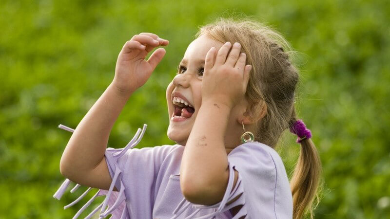 Lachendes blondes Mädchen mit lila Fetzenshirt, die Hände auf Höhe der Stirn