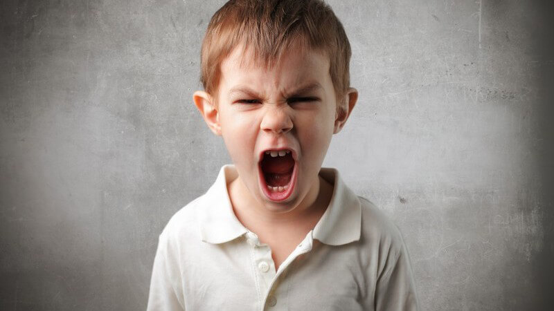 Kleiner Junge in weißem Poloshirt steht vor einer grauen Wand und schreit wütend