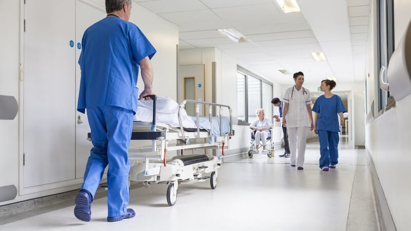 Krankenhausflur - Arzt schiebt Krankenbett, im Hintergrund Patienten, Krankenpfleger und Krankenschwester