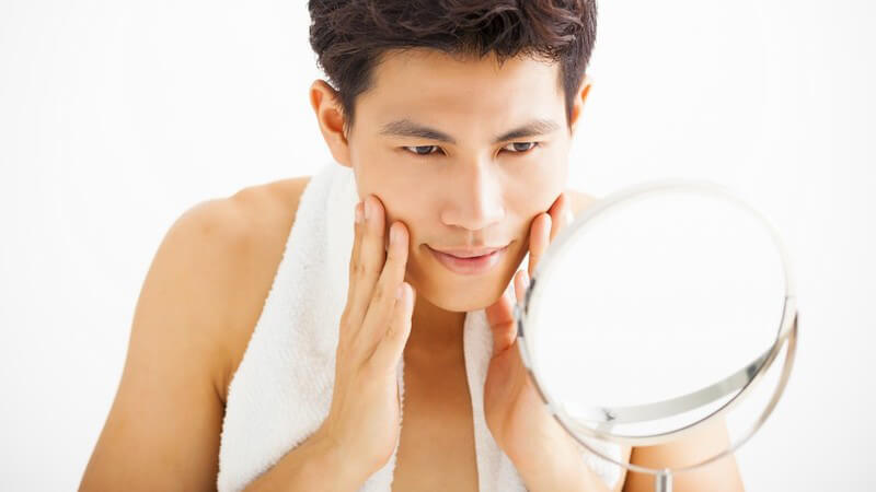 Junger asiatischer Mann mit Handtuch um den Hals betrachtet sich im Kosmetikspiegel und streicht sich über das Gesicht