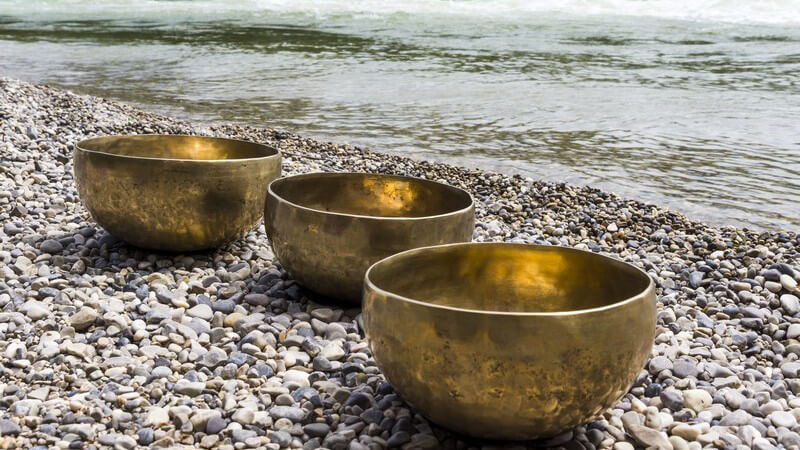 Drei goldene Klangschalen liegen auf Kieselsteinen am Wasser