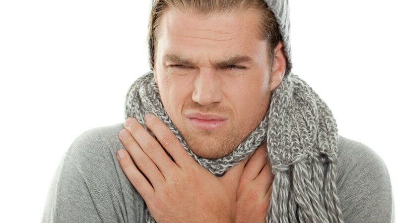 Halsschmerzen: junger Mann mit Schal und Strickmütze hält Hände an Hals