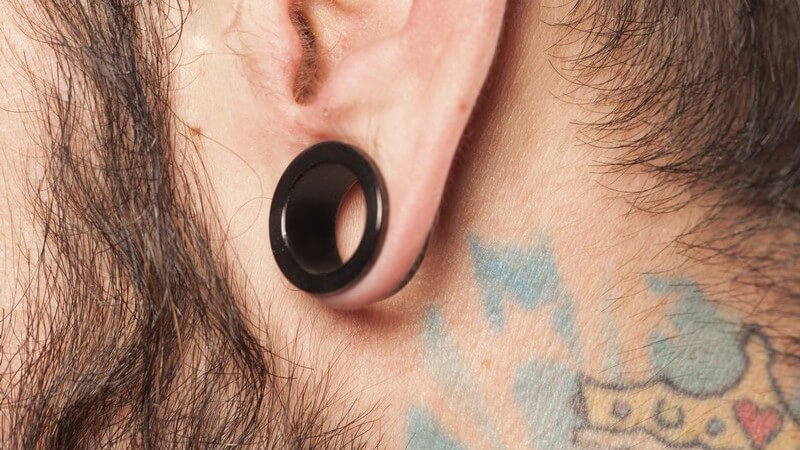 Gedehntes Lobe-Piercing (schwarzer Ohrring) am Ohr eines bärtigen Mannes mit Tattoo am Nacken