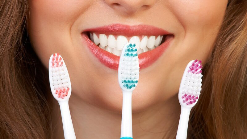 Lächelnde junge Frau hält drei Zahnbürsten vor ihr Gesicht