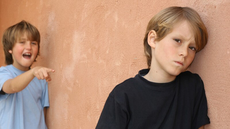 Trauriger Junge lehnt an Wand, hinter ihm zeigt Junge mit Finger auf ihn und lacht Ärgern Hänseln Mobbing Spott