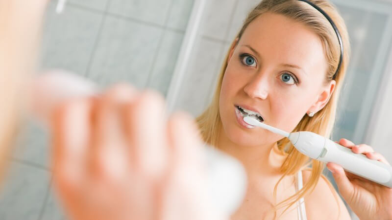 Blonde Frau mit schwarzem Haarband putzt sich vor dem Spiegel die Zähne mit elektrischer Zahnbürste