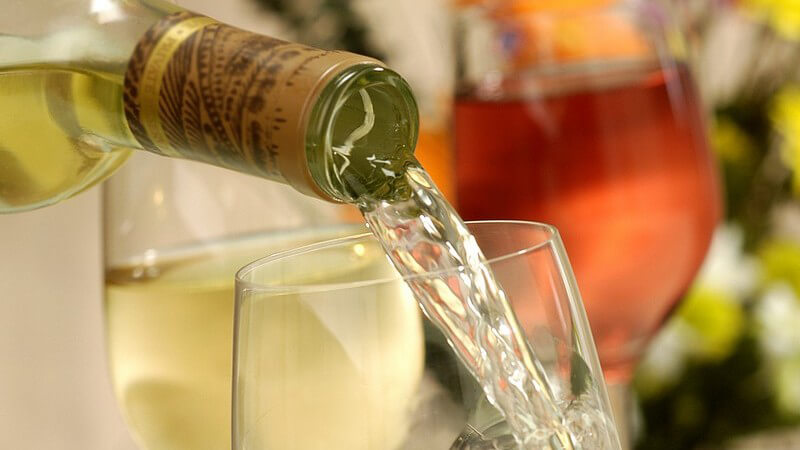 Weißweinflasche, die in Weinglas einschenkt, mit Weißweinglas und Rotweinglas im Hintergrund