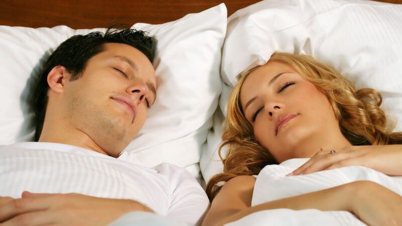 Links brünetter junger Mann mit kurzem Haar, rechts blonde Frau mit lockigen Haaren, schlafen in weißer Bettwäsche