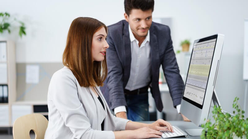 Junge Geschäftsfrau sitzt am Schreibtisch vor dem Computer, ein Kollege steht daneben und schaut mit auf den Monitor
