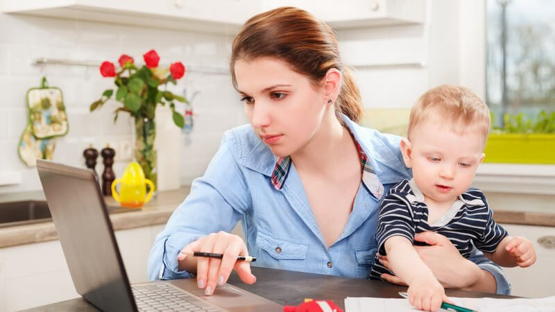 Junge Mutter sitzt in der Küche am Laptop und arbeitet, den Sohn auf dem Schoß