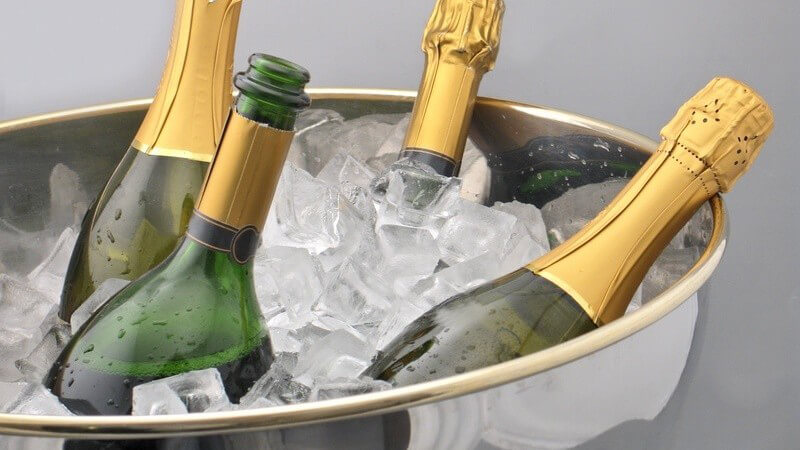 Vier Sekt- oder Champagnerflaschen in silbernem Sektkübel mit Eiswürfeln vor grauem Hintergrund