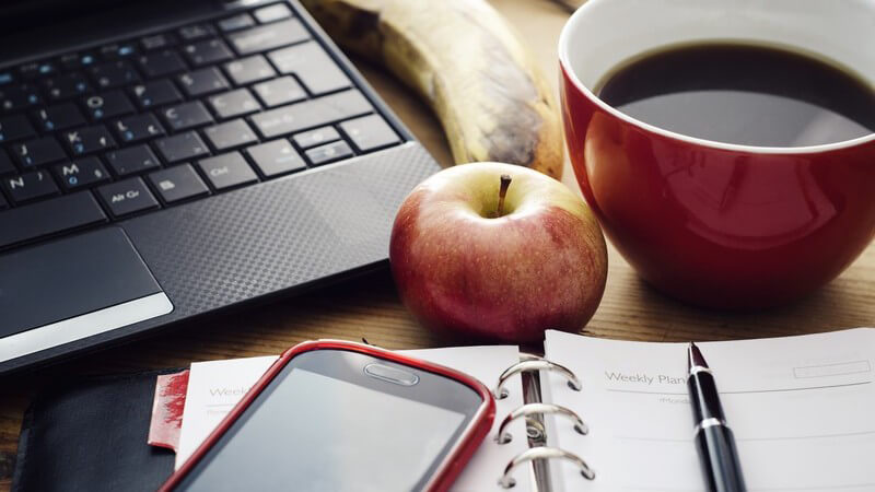 Arbeitsplatz mit Laptop, Handy, Notizblock, Kaffeetasse, Apfel und Banane