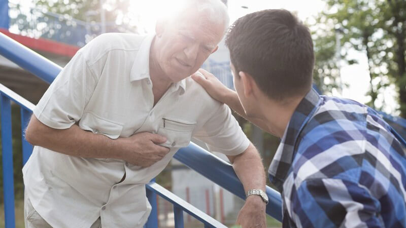 Alter Mann steht an einem blauen Treppengeländer und fasst sich ans Herz, ein junger Mann hilft ihm