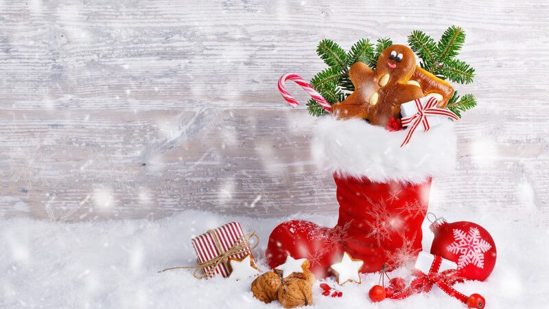 Roter Nikolausstiefel mit kleinen Geschenken und Süßigkeiten im Kunstschnee vor heller Holzwand