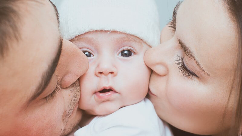 Säugling mit weißer Wollmütze wird von Mama und Papa geküsst