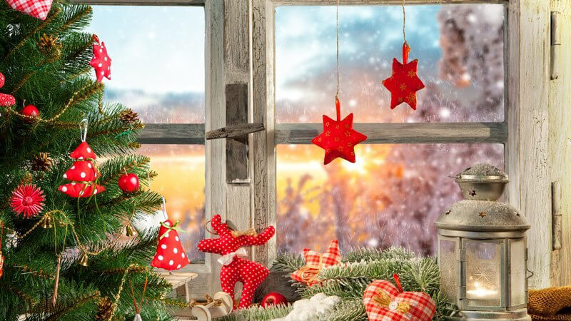 Weihnachtlich dekoriertes Holzfenster, Windlicht und rote Weihnachtsdeko, davor steht ein geschmückter Tannenbaum