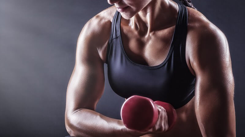 Muskulöse Frau in grauem Sport-BH beim Training mit roter Kurzhantel vor grauem Hintergrund