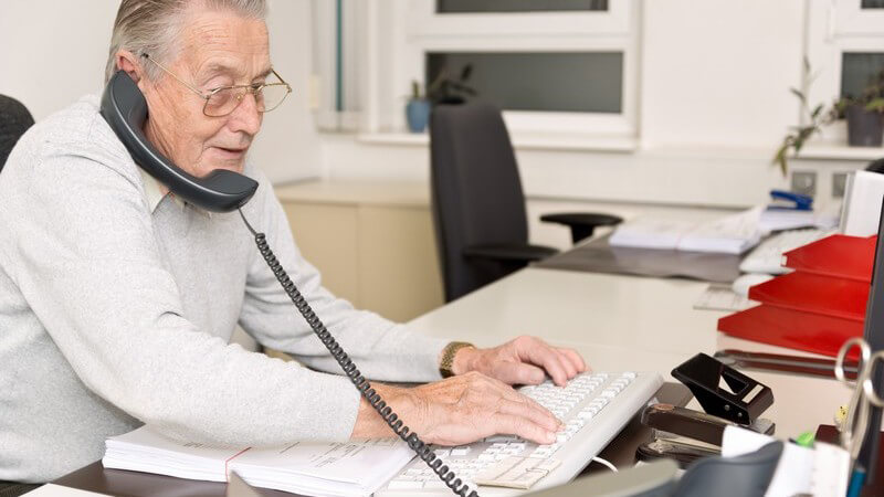 Alter Mann mit Brille sitzt im Büro am Schreibtisch und telefoniert