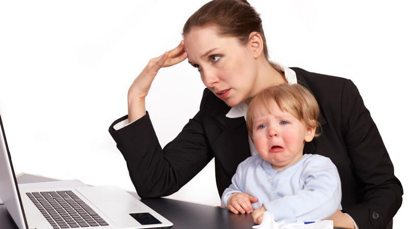 Gestresste Geschäftsfrau beim Arbeiten am Laptop, auf ihrem Schoß sitzt ein weinendes Kind