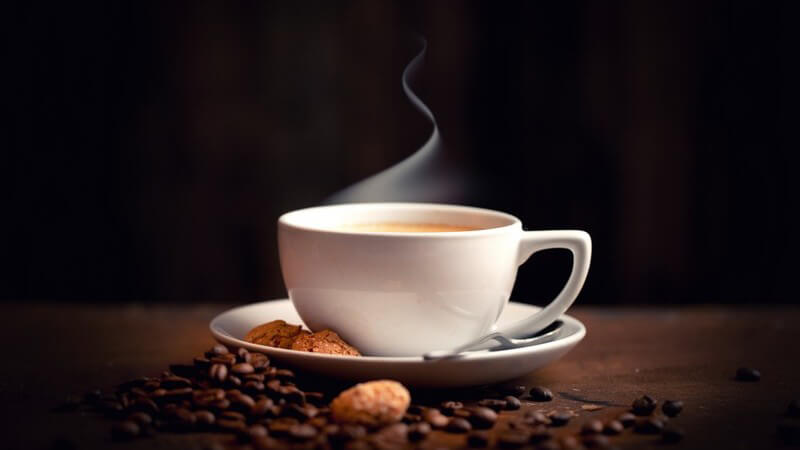 Dampfende Kaffeetasse neben kleinen Keksen und Kaffeebohnen vor schwarzem Hintergrund