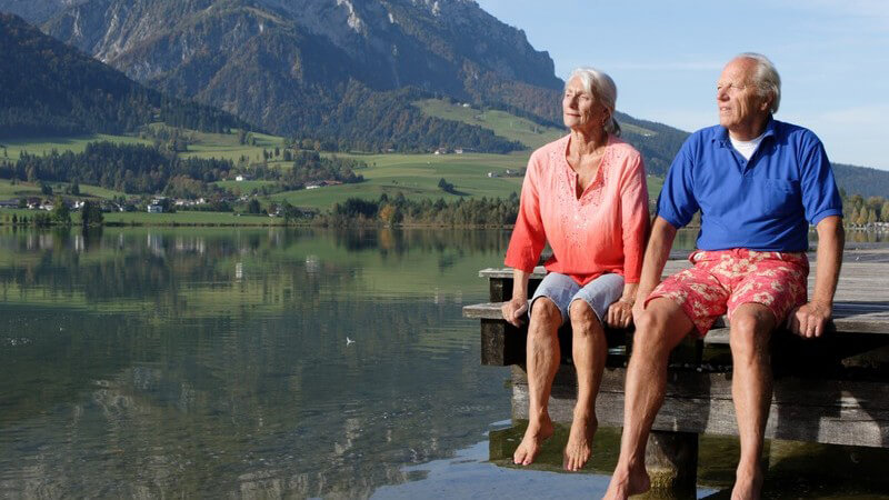 Älteres Paar sitzt auf Steg, nackte Füße über Wasser am Gebirgssee