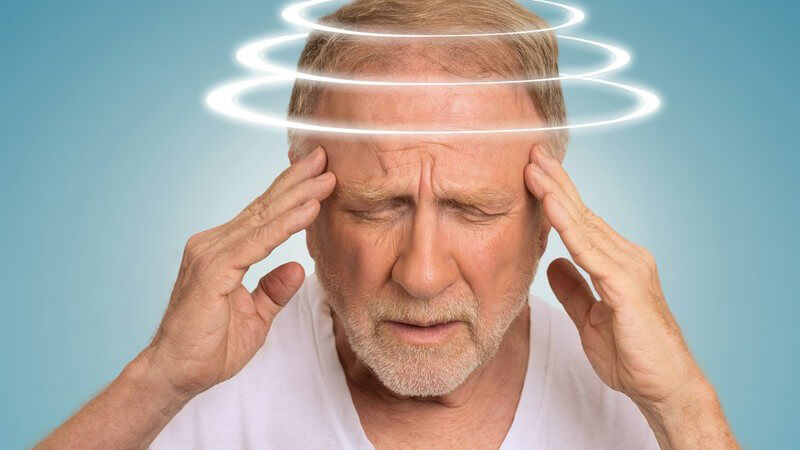 Senior in weißem Shirt hat Augen geschlossen und fasst sich an die Schläfen, drei Kreise schwirren um den Kopf