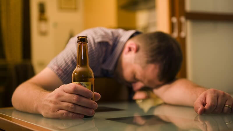 Betrunkener Mann über Tisch gebeugt, hält Flasche Bier in der Hand, Alkoholmissbrauch