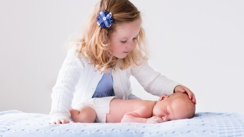 Kleines Mädchen in weißer Strickjacke streichelt ihrem neugeborenen Geschwisterchen über den Kopf