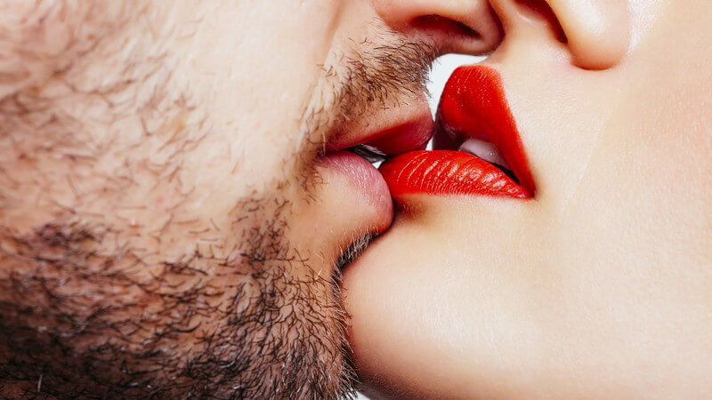 Mann mit kurzem Vollbart küsst die Unterlippe einer schönen Frau mit knallrot geschminkten Lippen