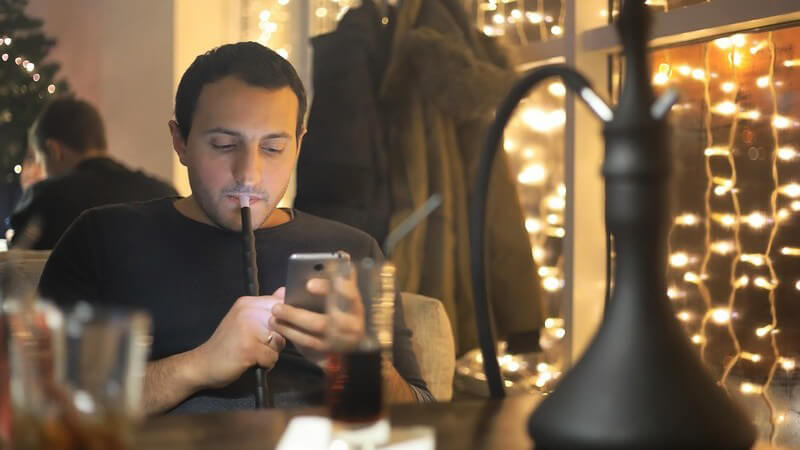 Mann sitzt in einer Bar an seinem Smartphone und raucht Wasserpfeife